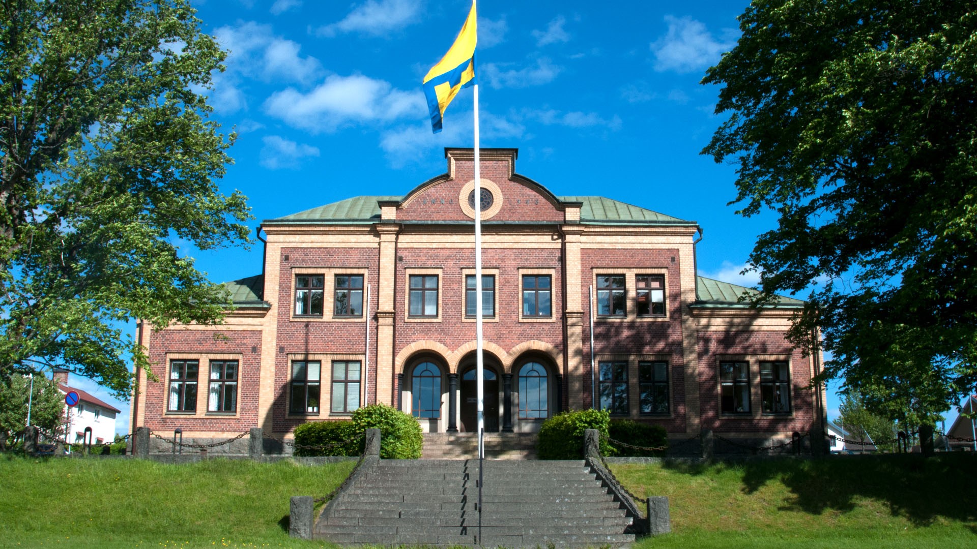 Bild på Sävsjö kommuns Kommunalhus  med Sävsjö kommuns flagga och logga framför. Bakgrund är en blå himmel med grönska. En trappa som leder upp till kommunalhuset finns med i bilden. 